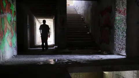 BBC A lone figure walks down a dark hallway in the ruined building, with graffiti on the walls