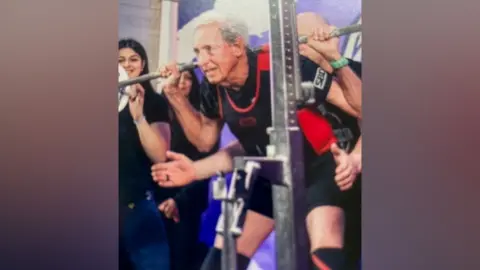 A man in his 80s holds a pair of weights on his shoulders as he lifts them from their holders. He is wearing black shorts, a black T-shirt, and a black and red vest. He has receding grey hair. Ther are women stood to his side, clapping on in support and a man stood behind him is hovering his hands over his hips for support, if needed.