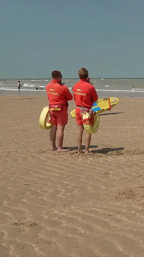 Two lifeguards on a sandy beach holding boards
