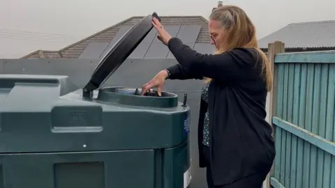 Ellen Knight/BBC Photograph of Sproson-Jones lifting up the lid of one of the oil tanks. She's wearing black trousers, a black cardigan, and a black floral blouse underneath. Her long blonde hair is tied back into a half-up hairstyle, and she's also wearing a pair of dark-framed glasses. The oil tank is large, coming up to about chest height, and is made of dark green plastic with a large black hatch which Sproson-Jones is lifting up. Behind it is a blue-painted fence, with some rooftops seen beyond the fence. The sky is grey and overcast. 