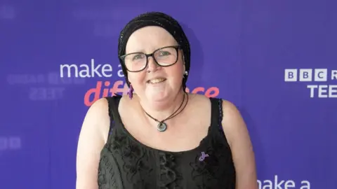 A woman in a black dress, headscarf and glasses smiles at the camera