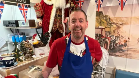 BBC/RICHARD EDWARDS Warren Mills - who runs a cafe in Filey with his wife - and is against the council tax premium - is looking straight at the camera. He has short dark hair and is wearing a Christmas T-shirt and a blue apron. There is a Father Christmas statue behind him, Union Flag bunting above him, and a painting of Filey's beach, with a fishing boat and tractor on it, to his right.
