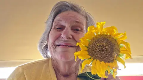 Cheri Gardiner Ann Holmes is 82 and has shoulder length white hair, she is smiling and is holding a sunflower to her left. She is pictured wearing a yellow cardigan and is a room with a mustard coloured wall.