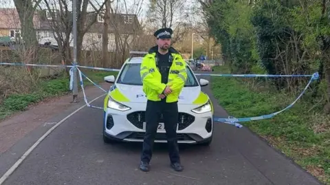 Essex Police A police officer wearing a hi-vis jacket stands on the cordoned-off cycle path. Behind him is a police car and blue and white tape. The path is lined by trees.