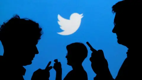 Reuters People holding mobile phones are silhouetted against a backdrop projected with the Twitter logo in this illustration picture taken in Warsaw September 27, 2013