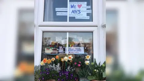 Farzana Chaudry Knitted figures in window with signs "Stay at home. Protect the NHS" and "We stayed at work for you now stay at home for us please"