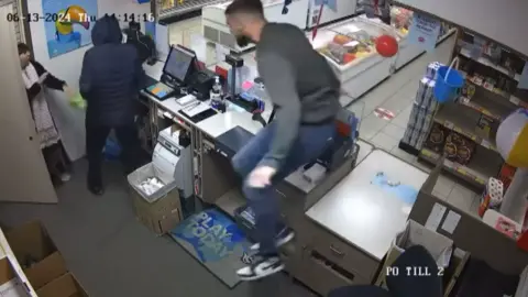 Usman Javid CCTV image of staff grabbing would-be robber Robert Daneskin behind the counter. One man is behind Daneskin and has his arms wrapped around him. Daneskin is wearing a balaclava and blue coat, while holding an imitation gun and a bag in his hand 