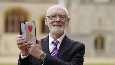 Martyn Butler with his OBE award in June 2022. He wears a dark suit with a pruple striped tie. 