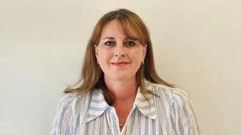 St Luke's Hospice A head and shoulders image of a woman with shoulder-length light brown hair, smiling at the camera and wearing a white and blue stripped casual shirt style top.
