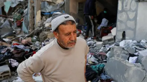 A man wearing a woolly hat and a grey jumper stands amidst debris in Khan Younis in southern Gaza Strip