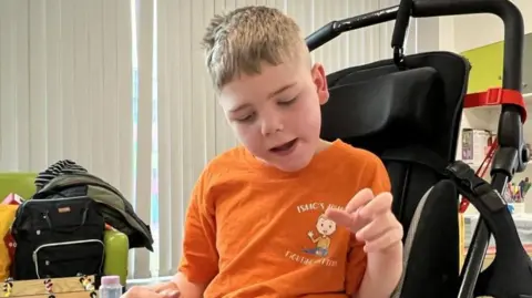 A boy with short blond hair in a pushchair wearing an orange t-shirt saying Isaac's Journey above a cartoon figure of a waving boy. The boy in the pushchair is looking downwards. Bags on a green sofa can be seen to his right, along with white vertical blinds. 