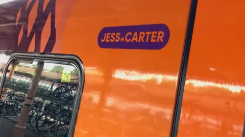 West Midlands Railway An orange and purple West Midlands Railway train with the name "Jess Carter" printed on in purple with a mini football in between her name.