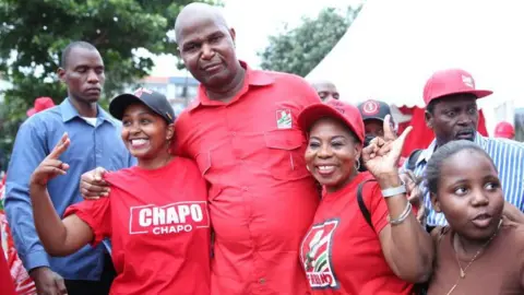 AFP Daniel Chapo celebrates his victory with supporters in Maputo in December. They are wearing red, the colour of his Frelimo party. Two woman put their arms around the politician and make the 'peace' sign with their hands.