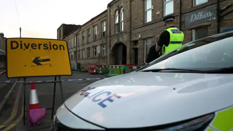 Mark Graham/BBC A street scene showing a police incident cordon outside a row of stone-built commercial and residential buildings. In the foreground, the bonnet of a marked police vehicle occupies the right side of the image, while a large yellow road sign on the left reads “Diversion” with a left-pointing arrow. A uniformed police officer wearing a high-visibility vest stands near the cordoned area, speaking with another person. Blue-and-white police tape and temporary road barriers block access to part of the street. In the background, a multi-storey stone building with an arched entrance is visible, along with a business sign reading “Dilbar’s.” The lighting suggests late afternoon or early evening, and the road appears closed as traffic is redirected away from the area.