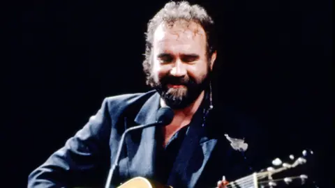 John Martyn treedt op op het podium - hij heeft een akoestische gitaar en draagt ​​een donker pak.