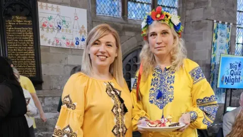 Two women with blonde hair wearing traditional yellow dress and one of them has a garland of flowers in her hair. They are standing in a church and one of them is holding a plate of food.