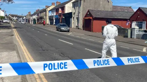 Blue and white police tape across a road, someone in a white forensics suit is walking up the road