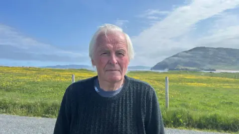 A man in a thick knitted jumper is standing in front of a field, filled with tall grass and small yellow flower heads. Behind him is a coastline and a mountain, with sprawling blue skies. The man has flowing grey hair and is looking directly at the camera. 
