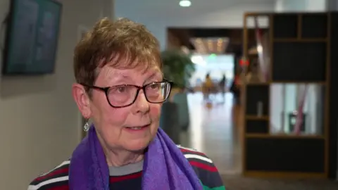 Lesley Mahmood has short, light brown hair. She wears dark-rimmed glasses, a striped jumper and a purple scarf