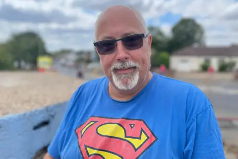 Laura Foster/BBC A man with black sunglasses and a blue T-shirt, with the red and yellow Superman logo on it, sits outside looking at the camera.