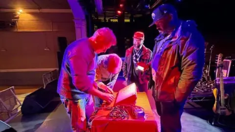 Carey Davis, Princess Pavilions Members of the band, Arrested Development, stood on a stage with a member of the Falmouth repair cafe who is fixing their sampler. The lighting is red and blue and the room is dimly lit, with a band member holding a flashlight.