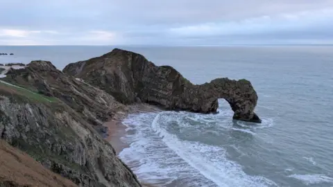 John Caines View from the top of the cliffs of Durdle Door. The dark limestone sea arch sticks out from the cliffs in a rough, grey sea on a cloudy day.