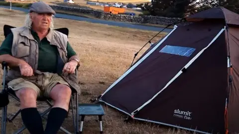 Wander with Mac/YouTube Man sitting on a camping chair next to a tent