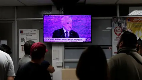 Reuters People watch a television broadcasting a speech by Chile's President Sebastian Pinera in an emergency room of a hospital amid anti-government protests in Santiago, Chile, 12 November, 2019