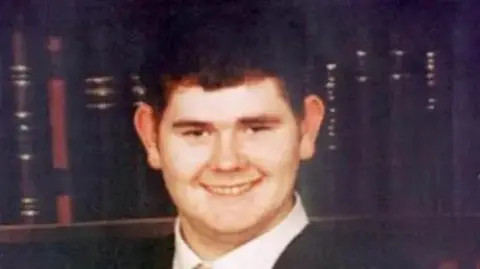 The family handout Aiden Gallagher smiles as he stands in front of a book wardrobe wearing a black suit or school blazer and white shirt. he has short black hair.