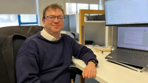 Martyn Dorey - A man with wavy brown hair and glasses leaning on a white desk. He's wearing a blue Guernsey jumper over a white shirt. 