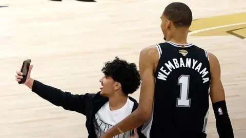 A fan runs on to the court and takes a photo with Victor Wembanyama during game one of the 2026 NBA Finals