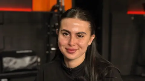 Rhianna Venables/BBC A girl with straight dark brown hair smiling at the camera. She has a silver nose ring on her left side.