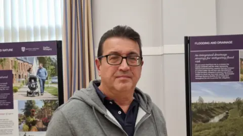 Simon Bradshaw A man in a hoodie. He has dark hair and glasses and is standing in front of boards showing a planning application