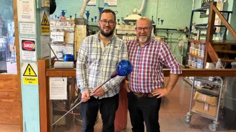Two men wearing checkered shirts stand side-by-side. One holds a bit of Bristol Blue Glass. There is a glassware factory in the background. 
