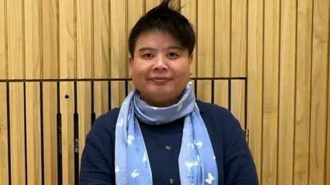 Vicky Makaprang standing in front of a wooden panel, looking at the camera and wearing a navy cardigan and light blue scarf.