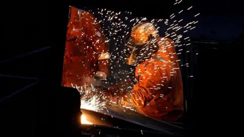 Sparks fly as workers wearing hi-vis orange jackets and helmets work with metal at a steel factory in Scunthorpe.