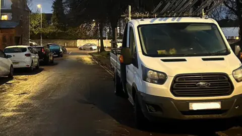 A broken into white work truck on a residential street, the door has been drilled through. It's dark outside and the street is dimly lit. 
