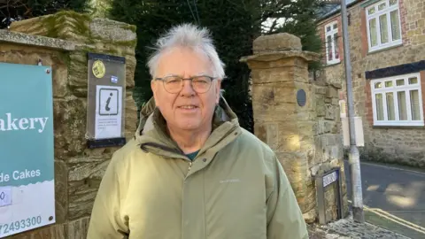 A white man with grey hair in front of a cream stone wall