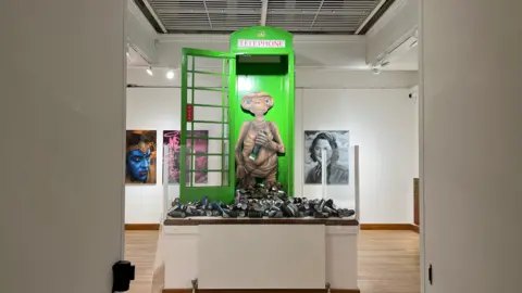 Tom Jackson/BBC A setting inside the museum- with paintings on a white wall in the background. the focus of the pic is a green telephone box with many spary cans left in front of it and a cut-out of a yodha like character standing inside it. 