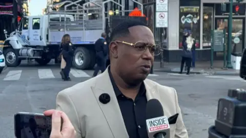 Rap singer Master P speaks to reporters in New Orleans' French Quarter. He wears gold-rimmed glasses, a black shirt and beige suit jacket. A microphone is being held in front of his face.