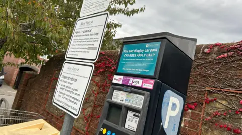 LDRS A New Forest District Council car park ticket machine in front of a brick wall with white regulations signs to its left