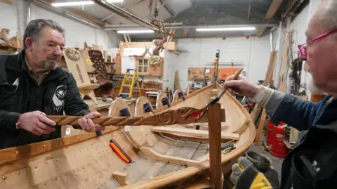 Owen Humphries/PA Two men are painting a piece of wood carved into the shape of a dragon on either side of a Viking boat. They are both older men in their 60s or 70s.