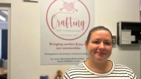 A woman with dark hair tied back in a striped white and black t-shirt stands in front of a white wall with a sign in the background. The sign has pink lettering saying Crafting and a logo of a balled up woollen heart with knitting needles crossed through it.