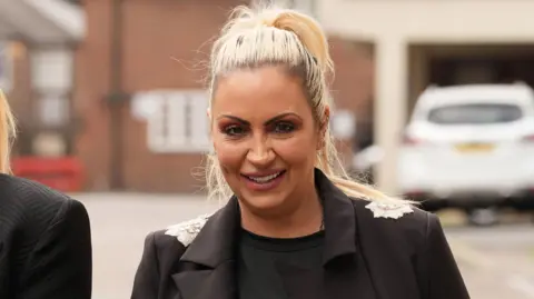PA Media Jodie Marsh has blonde hair in a high pony tail. She is wearing a black top and a black blazer with silver detail on the shoulders. Behind her is a building and a parked white car. 
