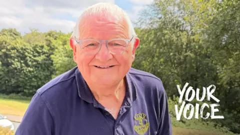 A man wearing a navy blue polo shirt wearing glasses looks relaxed and is smiling as he stands in front of a tree-lined street.
