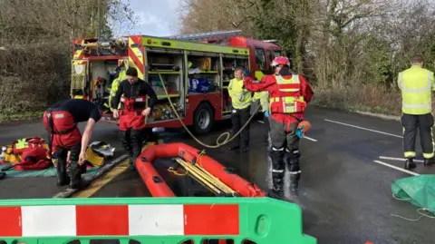 A number of firefighters put on red wet weather clothing next to a fire engine in a carpark. The are in the process of inflating a small boat. There us a green plastic piece of fencing near the front of the image.