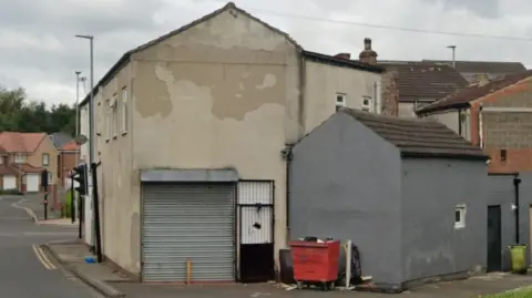 Google Street view of a decrepit looking two-storey building, whose cream coloured rendering has chipped away. There is a white door with a black metal grate across it, and a grey garage door.
