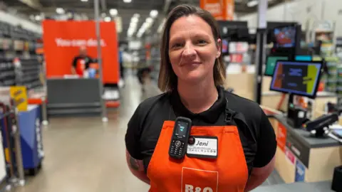 BBC News A female employee stands by a till in a B&Q store. She has shoulder length brown hair, and is smiling. She's wearing a black polo shirt with an orange apron over the top. She has a phone attached to it, and her name badge says 'Jeni'.