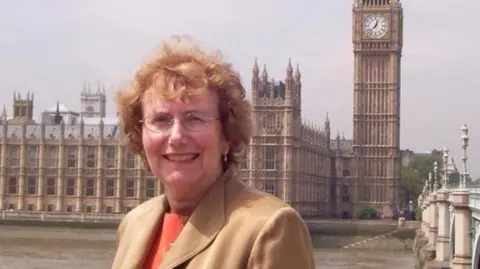 BBC An older picture of Dame Annette standing in front of the Houses of Parliament. She has red hair and wears a beige coat and orange top.