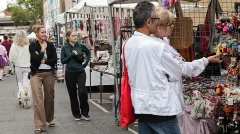 Getty Images A busy street market. On front right of picture, a man and a woman are looking at key rings with stuffed animals on them. To their left and further away, two women are looking at a stall with pictures on it. Behind them is a woman with a dog and there are other people are stalls further in the background.